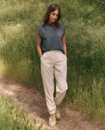 A woman on a grassy path embodies outdoor spirit, wearing a sleeveless polka dot shirt, THE GREAT. SP25 TGO Trailhead Pant in cream, and beige boots, hands casually tucked in pockets.