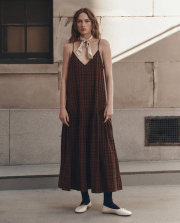 A woman with wavy hair wears THE GREAT. F26 D1's The Drop Waist Dress in Woodcrest Plaid—a long brown plaid dress with spaghetti straps—paired with a white neck scarf, dark leggings, and white ballet flats as she stands in front of a stone building.
