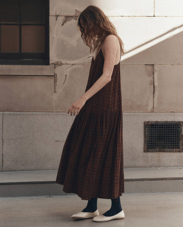 Wearing THE GREAT. F26 D1's "The Drop Waist Dress" in Woodcrest Plaid, a person with long hair stands beside a textured building wall, paired with dark socks and light-colored flat shoes.