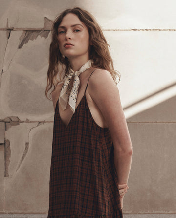 A woman with wavy hair wears THE GREAT. F26 D1 The Drop Waist Dress in Woodcrest Plaid and a light patterned scarf around her neck, standing against a textured wall.