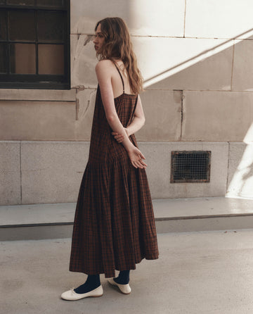 A woman with wavy hair stands outside a stone building wearing THE GREAT. F26 D1's The Drop Waist Dress in Woodcrest Plaid, paired with dark socks and white ballet flats.