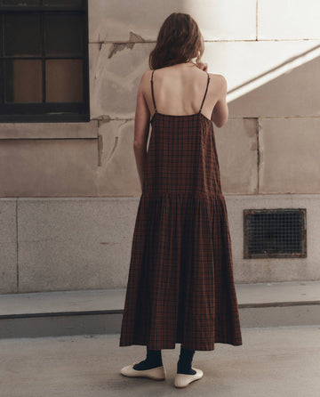 A woman with long hair stands outdoors facing a wall, wearing THE GREAT.'s The Drop Waist Dress in Woodcrest Plaid, paired with black socks and white shoes.