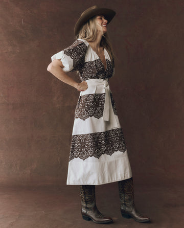 A woman in profile wears THE GREAT. F25 D1's The Fairground Dress in cream with western lace print, styled with a wide-brimmed hat and cowboy boots, set against a plain brown backdrop.