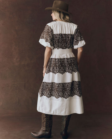 A person wears THE GREAT. F25 D1 The Fairground Dress—Cream With Western Lace Print—paired with a brown wide-brimmed hat and cowboy boots, standing against a brown textured background.