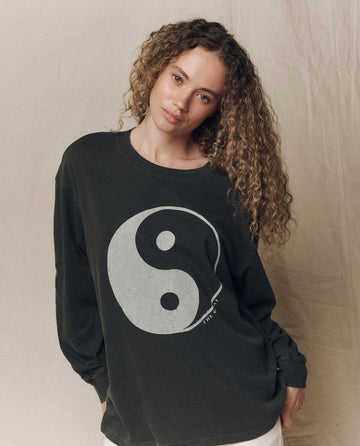 A person with curly hair wears THE GREAT. F26 HALLOWEEN GRAPHICS “The Long Sleeve Relaxed Crew w/ Yin Yang Graphic” in washed black, standing against a plain beige background.