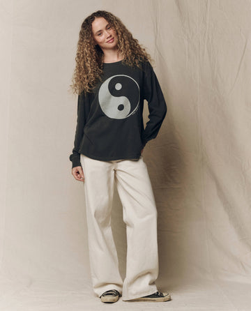 A person with curly hair wears THE GREAT. F26 HALLOWEEN GRAPHICS The Long Sleeve Relaxed Crew w/ Yin Yang Graphic in washed black, paired with cream wide-leg pants and black sneakers, standing against a beige fabric backdrop.