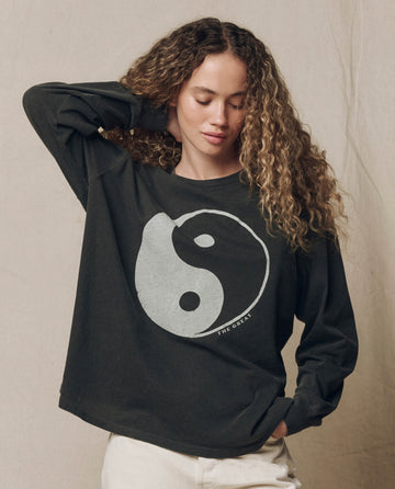A person with curly hair wears THE GREAT. F26 HALLOWEEN GRAPHICS The Long Sleeve Relaxed Crew w/ Yin Yang Graphic in Washed Black, paired with light-colored pants, standing against a plain beige background.
