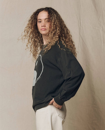 A person with long, curly hair stands sideways against a neutral, textured backdrop wearing THE GREAT.’s The Long Sleeve Relaxed Crew w/ Yin Yang Graphic in Washed Black, part of the F26 HALLOWEEN GRAPHICS collection, paired with light pants.