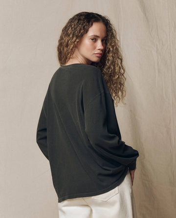A person with long curly hair stands in front of a neutral fabric backdrop, looking over their shoulder while wearing THE GREAT. F26 HALLOWEEN GRAPHICS The Long Sleeve Relaxed Crew w/ Yin Yang Graphic in Washed Black, paired with light pants.
