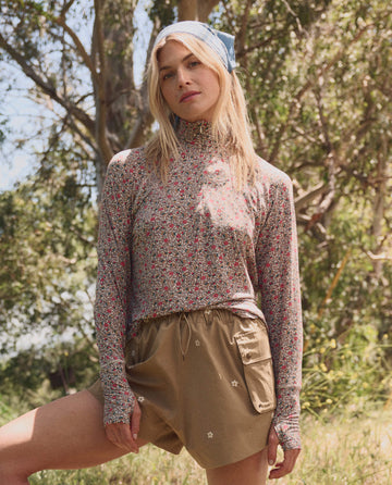 A woman in THE GREAT. SP26 TGO The Quarter Zip Trail Pullover – Sprouting Floral, with khaki shorts and a blue headscarf, stands outdoors by trees in daylight.