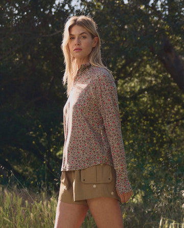 A woman stands outdoors in sunlight wearing THE GREAT. SP26 TGO The Quarter Zip Trail Pullover – Sprouting Floral and tan shorts, surrounded by trees and grass.