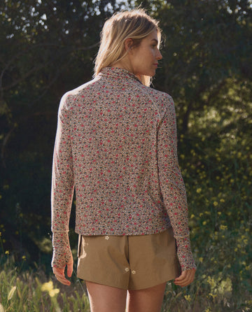 A person stands outdoors facing away, wearing THE GREAT. SP26 TGO The Quarter Zip Trail Pullover in Sprouting Floral and tan shorts, surrounded by greenery as sunlight filters through the trees.