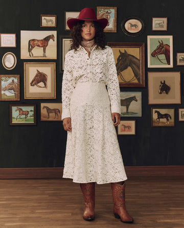 Woman in a white Western dress styled with THE GREAT. F26 STETSON’s The Relic Lace Saddle Skirt in cream, a red cowboy hat, and brown boots stands indoors against a dark wall adorned with framed horse artwork, capturing the essence of classic American craftsmanship.