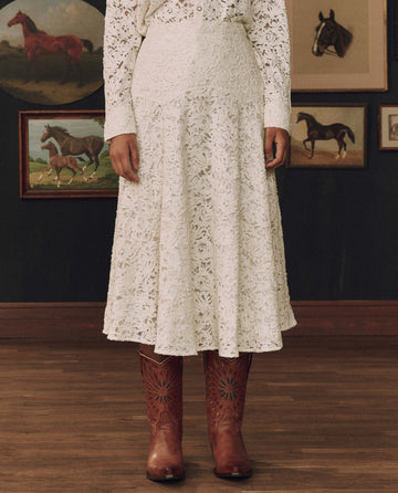 A person wears THE GREAT. F26 STETSON’s The Relic Lace Saddle Skirt in cream—a long, intricately laced Western piece inspired by American craftsmanship—standing indoors before paintings of horses and finishing the look with brown cowboy boots.