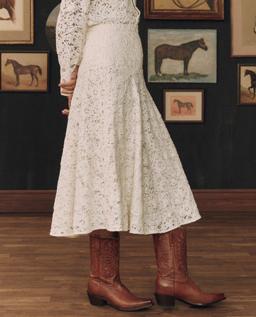 A person stands indoors wearing THE GREAT. F26 STETSON’s The Relic Lace Saddle Skirt in cream, styled as a white lace Western dress with brown cowboy boots. Behind them, framed horse artwork decorates the dark wall—an homage to timeless American craftsmanship.