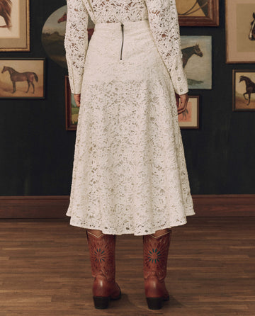 A person stands indoors on a wooden floor, facing away from the camera, wearing THE GREAT. F26 STETSON’s The Relic Lace Saddle Skirt in cream and brown cowboy boots. Framed horse images on the dark wall in the background evoke a sense of American craftsmanship and Western style.