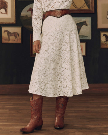 Dressed in THE GREAT. F26 STETSON’s The Relic Lace Saddle Skirt in Cream, cinched with a brown tooled leather belt and paired with classic brown cowboy boots, this individual stands on a wooden floor against a backdrop of horse paintings—embodying the spirit of Western dress and true American craftsmanship.