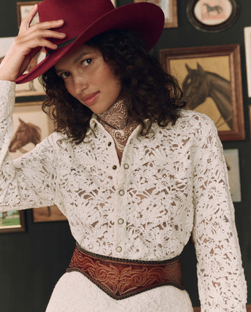 A woman wearing a lace white blouse, a vibrant red THE GREAT. F26 STETSON cowboy hat, and The Tooled Corset Belt in Burnished Brown stands in front of a wall adorned with framed horse art.