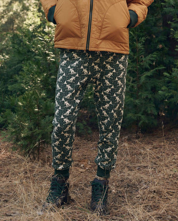 Person standing outdoors in a brown puffer, THE GREAT. F26 TGO The Union Long John. -- Spruce with white animal shapes, and green hiking boots among trees and dry grass.