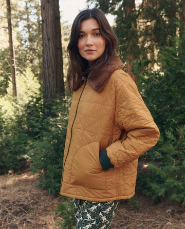 A person wears THE GREAT. F26 TGO The Reversible Logger Puffer. -- Pine And Amber, standing outdoors in a forested area with patterned pants.