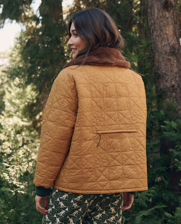 A woman wears The Reversible Logger Puffer in Pine And Amber, a mustard yellow reversible puffer jacket with a brown faux-fur collar and a horizontal zippered pocket on the back.