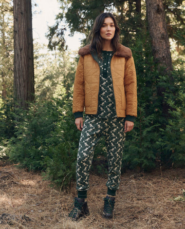 A person in The Union Long John. -- Spruce, a quilted mustard-colored jacket with a faux fur collar, and hiking boots in a forest.