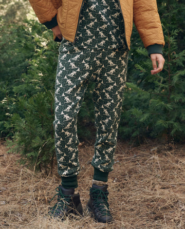 Person standing outdoors in The Union Long John. -- Spruce by THE GREAT. F26 TGO, with a brown quilted jacket, green sweater, dark hiking boots; green bushes and dry grass behind.