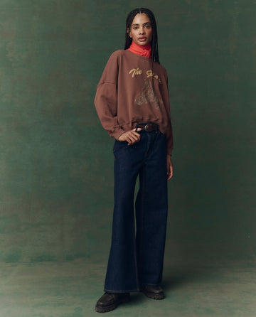 A person stands on a green background, wearing THE GREAT. F25 D2’s The Teammate Sweatshirt—Umber with Horse Embroidery—paired with a red turtleneck, wide-leg dark jeans, and black shoes.