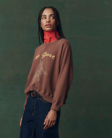 A woman wearing THE GREAT. F25 D2's The Teammate Sweatshirt in umber with horse embroidery, paired with dark jeans and a red turtleneck, stands against a green background, gazing to the side.