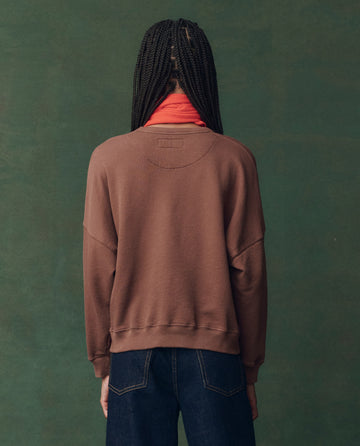 Person with braided hair wears THE GREAT. F25 D2 The Teammate Sweatshirt in umber with horse embroidery, red turtleneck, and dark jeans, standing against a green background with their back to the camera.