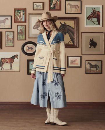 A woman in a beige Stetson SP26 cowboy hat and THE GREAT. Western Riding Cardigan Sweater in Robin Blue stands indoors before a wall adorned with horse paintings, wearing a blue embroidered skirt.