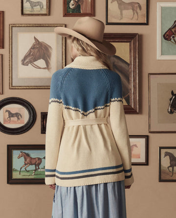 Wearing THE GREAT. STETSON SP26’s The Western Riding Cardigan Sweater in Robin Blue, a person in a wide-brimmed hat stands before a wall adorned with framed horse paintings.