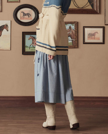 A person in a blue skirt, cream sweater, and THE GREAT. STETSON SP26 The Cowboy Boot (Suede) – Pearl stands indoors beside a wall adorned with framed horse paintings.