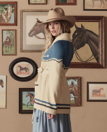 Wearing THE GREAT. STETSON SP26’s Western Riding Cardigan Sweater in Robin Blue, a woman in a cowboy hat stands before a wall of framed horse paintings.