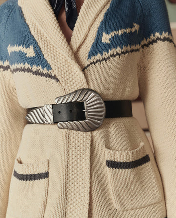A person wearing THE GREAT. STETSON SP26 Western Riding Cardigan Sweater in Robin Blue, styled with front pockets and cinched at the waist with a wide black belt featuring a large, textured silver buckle.