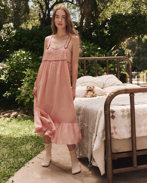 A woman wears THE GREAT. SP25 SLEEP's The Western Tank Night Dress in Rose Bloom, standing by a metal-framed bed outdoors with greenery and sunlight, while a teddy bear sits on the bed.
