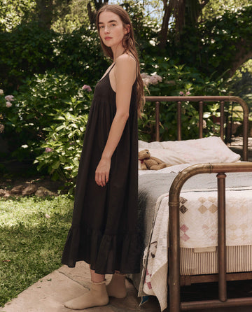 A woman in THE GREAT. SP25 SLEEP's Eyelet Ruffle Tank Night Dress in black cotton voile stands outdoors by a vintage metal bed with a quilt, surrounded by greenery.