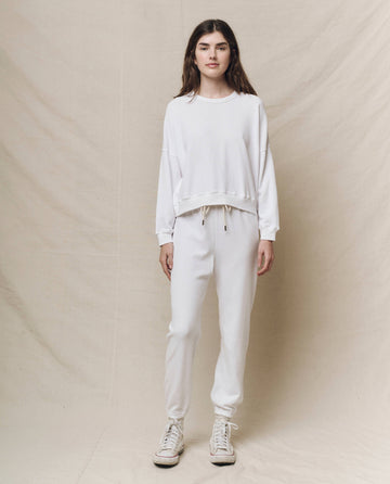 A person stands against a beige backdrop, wearing THE GREAT. HOL 24 KNITS' The Teammate Sweatshirt in True White, paired with vintage sweatpants and white sneakers, capturing the cool handcrafted Los Angeles style.