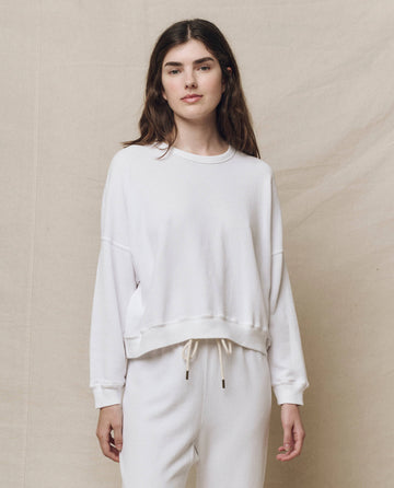 A woman in The Teammate Sweatshirt by THE GREAT. HOL 24 KNITS and white pants stands against a beige backdrop, showcasing timeless style with handcrafted Los Angeles flair.
