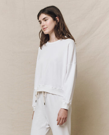 Against a beige backdrop, a person is styled in The Teammate Sweatshirt in True White and matching pants from THE GREAT. HOL 24 KNITS, exuding effortless Los Angeles flair.