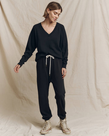 A person stands in front of a beige backdrop wearing The V-Neck Sweatshirt in Almost Black by THE GREAT. FALL 24 KNITS, paired with black pants featuring white drawstrings and white sneakers.