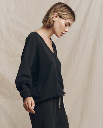 A person with short hair wearing THE GREAT. FALL 24 KNITS' "The V-Neck Sweatshirt" in Almost Black and matching black pants stands against a beige fabric backdrop. They are looking down with a composed expression.