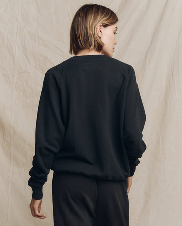A person with short hair is standing with their back to the camera, wearing a dark Almost Black V-Neck Sweatshirt by THE GREAT. FALL 24 KNITS and pants. They are against a plain, light-colored backdrop.