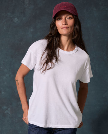 Woman wears The Boxy Crew. Solid -- True White by THE GREAT. CORE KNITS, blue jeans, and a burgundy cap before a dark textured background.