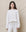 A person stands against a beige background wearing The Slouch Sweatshirt in True White by THE GREAT. HOL 24 KNITS, paired with matching pants, giving off a handcrafted vintage vibe, with their hands casually by their sides.