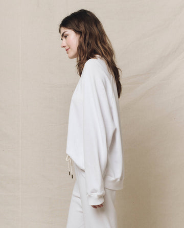 A woman in a relaxed white ensemble, wearing THE GREAT.'s "The Slouch Sweatshirt" in True White, stands sideways against a simple fabric backdrop.