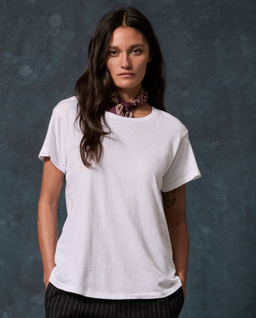 A woman with long dark hair wears The Slim Tee. Solid -- True White by THE GREAT. CORE KNITS, paired with a patterned scarf and striped pants, standing hands-in-pockets.