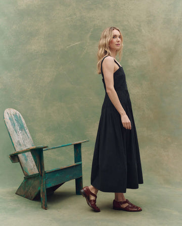 A woman in THE GREAT. SU25 The Day Break Dress -- Black and brown shoes stands by a weathered green wooden chair against a textured greenish background, embodying timeless fashion elegance.