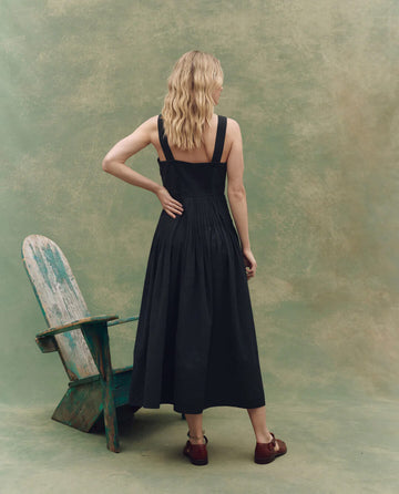 A woman with wavy blonde hair faces away, wearing THE GREAT. SU25's The Day Break Dress in Black and brown shoes by a weathered wooden chair, set against a textured backdrop for a timeless fashion statement.