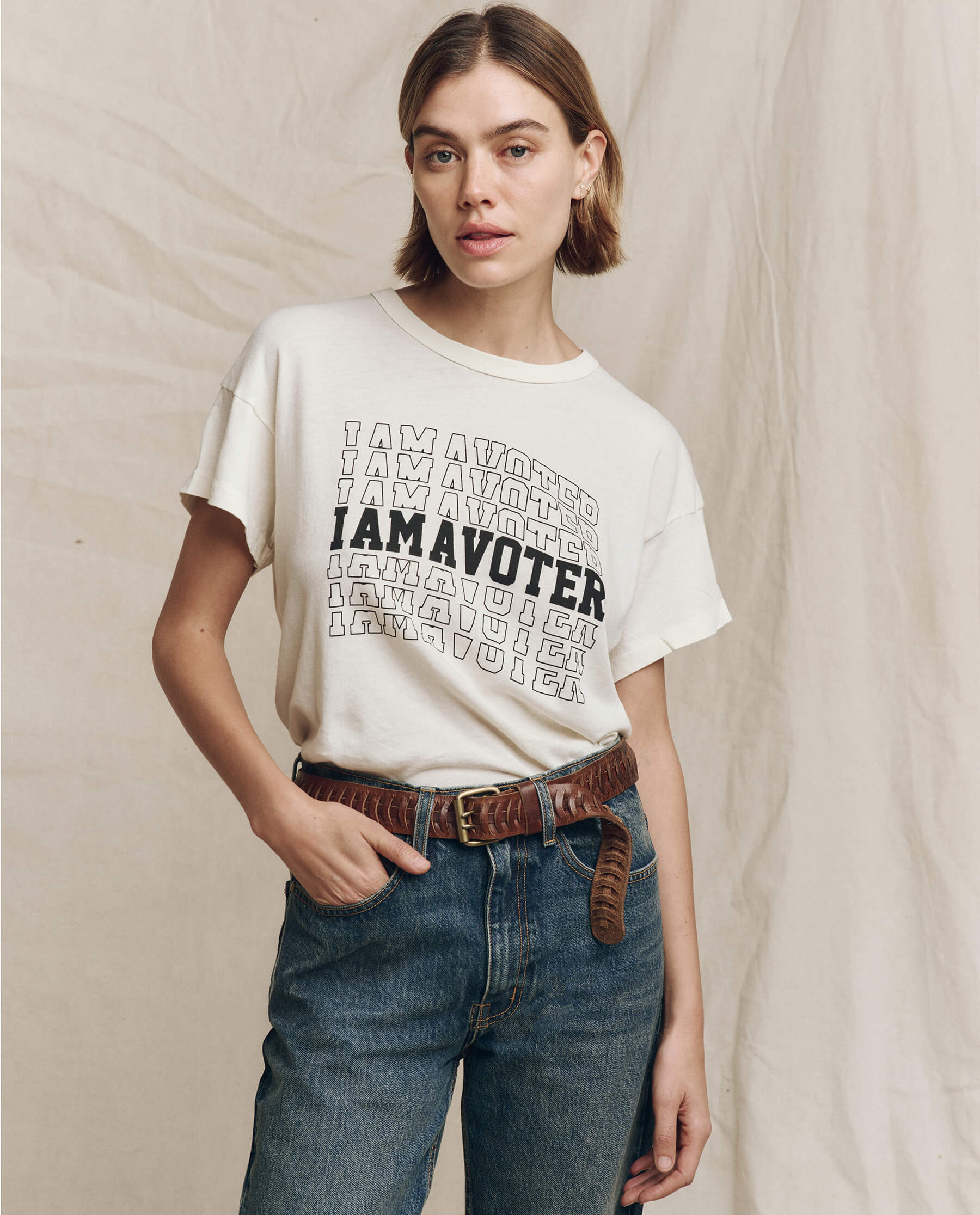 A person wearing a washed white "Limited Edition: The I Am A Voter Boxy Crew" tee by The Great. and jeans, standing against a neutral fabric backdrop.